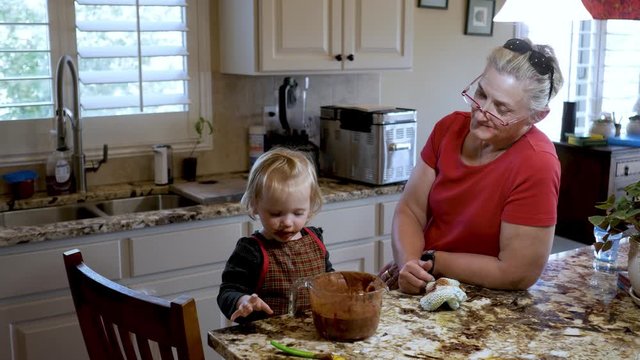 Grandmother makes chocolate pudding and toddler granddaughter eats it from the bowl