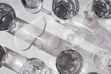 Horizontal close up of many glasses of water on white surface and dropped wineglass with spilled water with shadows on sunlight of sunset or sunrise.Drinking fresh water for health and beauty concept