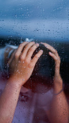 Fototapeta premium Person with a mask on his face while looking out the window with water drops.