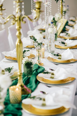 banquet table is decorated with a tablecloth, candles, compositions of greenery, on the table there are golden plates with napkins