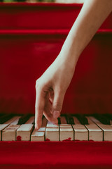 a close-up of a side view with female hand with pink manicure playing the piano. An old red piano with black and white keys. A palm above the keyboard. Graceful fingers of composer or musician
