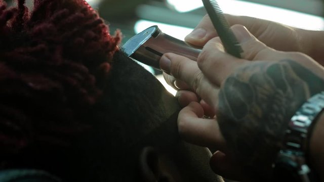 Close-up: a hairdresser shaves off the hair on the head of an African American. Makes a stylish hairstyle, uses an electric shaving trimer. Dreadlocks are red on the head