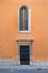 Fototapeta na wymiar Italy, Rome - February 24, 2012: .Ancient orange building in Rome. The big black door is closed, a huge window is located above it. The street is empty.