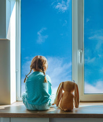 Little girl sitting back on the windowsill with bunny soft toy looking outside