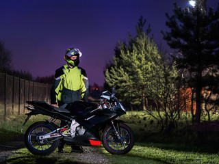 Fototapeta na wymiar A sports bike is on the road.A young man stands nearby.In a helmet. On one side is a chain-link fence and on the other are trees. Sunset. The sky is blue and purple. Motorcycle