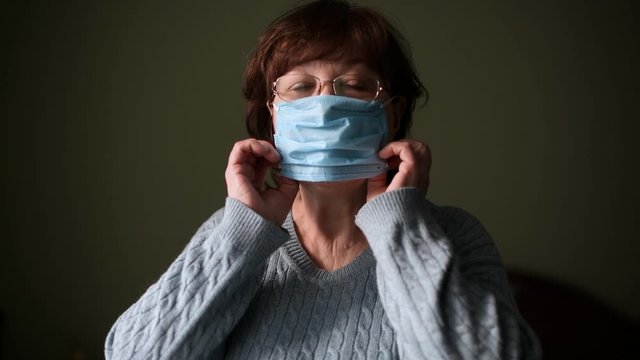 An aged woman puts on a protective medical mask on a dark background and then removes it, waving with relief