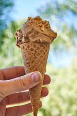 Hand holding an ice cream cone with peanut butter banana flavor and sprinkles of peanuts on top on a sunny day - gluten-free waffle cone 