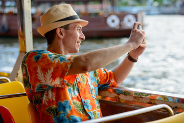 Fototapeta premium Happy young tourist man taking picture with phone while riding boat on the river