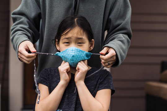 Adult assists Asian girl tie on cloth mask