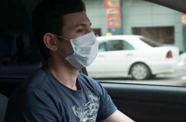 Portrait of man wearing disposable medical facemask in a car during coronavirus outbreak.Young man in protective sterile medical mask driving car. 