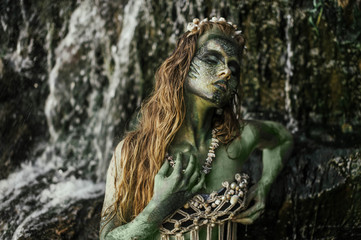 Fototapeta na wymiar girl mermaid amphibian with green skin with body art with a necklace and a seashell swimsuit around her neck holding her hand to her serious face in the river summer in the water in the lake