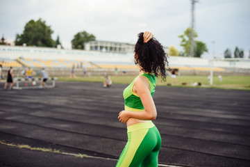 Beautiful curly girl flexing muscles in the stadium. The girl goes in for sports. Light green tracksuit