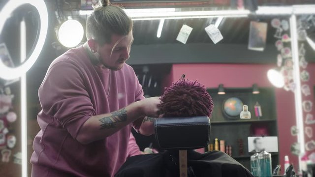 Close-up: a hairdresser shaves off the hair on the head of an African American. Makes a stylish hairstyle, uses an electric shaving trimer. Dreadlocks are red on the head