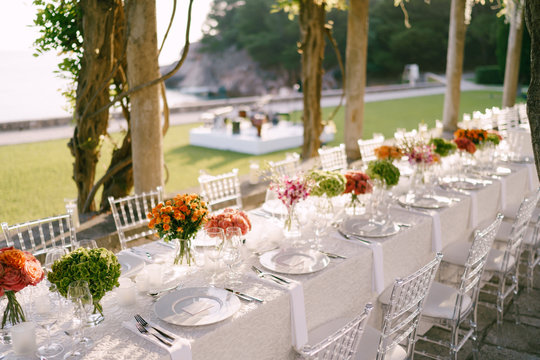 Wedding Dinner Table Reception. A Very Long Table For Guests With A White Tablecloth, Floral Arrangements, Glass Plastic Transparent Chairs Chiavari. Under The Old Columns With Vines Of Wisteria.