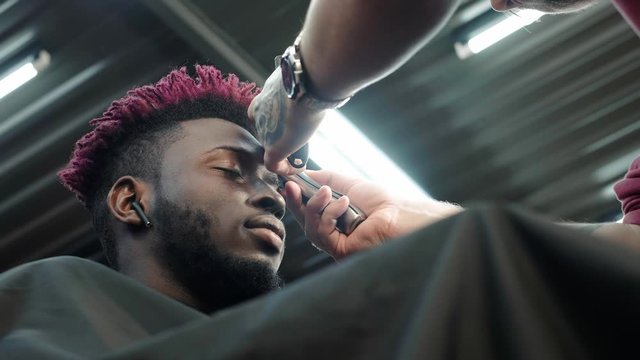 Close-up: a hairdresser shaves off the hair on the head of an African American. Makes a stylish hairstyle, uses an electric shaving trimer. Dreadlocks are red on the head