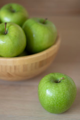 Green apples in a wooden bowl and one green Apple lying next to the bowl on the table.