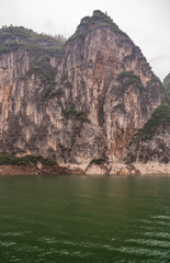 Baidicheng, China - May 7, 2010: Qutang Gorge on Yangtze River. Straigth down brown cliff with some green foliage on top and darker patches above green water.