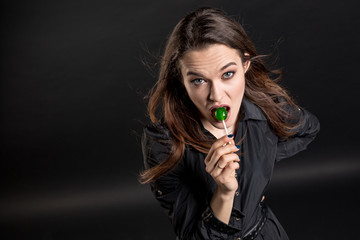 A makeup girl brunette in dark clothes licks a green candy on a stick lollipop and looks at the viewer. Studio portrait on a dark background. Horizontal orientation. 