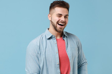 Cheerful funny young bearded guy 20s in casual shirt posing isolated on pastel blue background studio portrait. People sincere emotions, lifestyle concept. Mock up copy space. Blinking looking camera.
