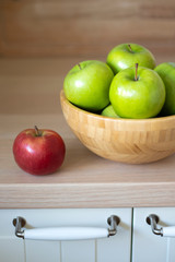 Green apples in a wooden bowl and one red Apple separately on the kitchen table. You can see the handles of the kitchen table doors.