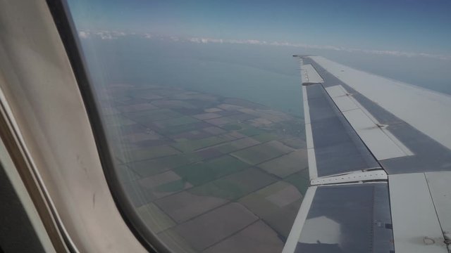 The plane flies over beautiful colorful fields, view from the porthole. Airplane wing in flight over fields, water, sea