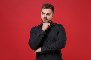 Handsome young bearded guy 20s in classic black shirt posing isolated on red wall background studio portrait. People lifestyle concept. Mock up copy space. Put hand prop up on chin, looking aside.