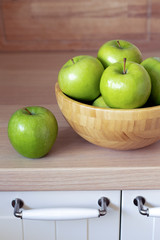 Green apples in a wooden bowl and one green Apple separately on the kitchen table. You can see the...