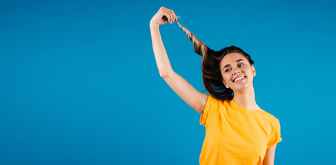 Close up of a smiling young woman with beautiful hair, holding hair with hands on blue background