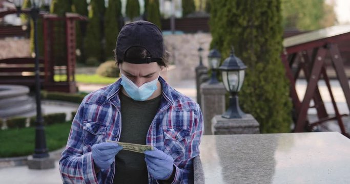 Man in mask and gloves holds last dollar in hands, looks on it, shows at camera