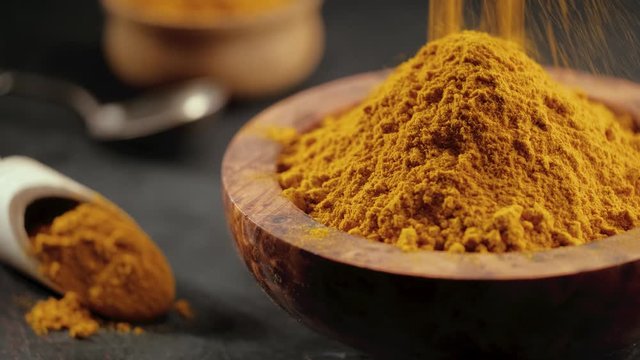 Turmeric powder or curcuma powder falling in a wooden bowl on a dark background, close-up. Cooking ingredients, flavor.