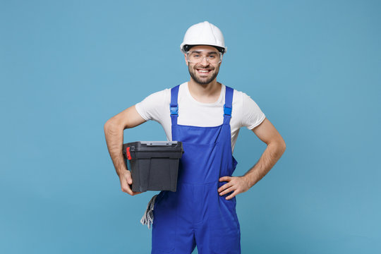 Smiling man in coveralls helmet hardhat glasses hold tool case box isolated on blue background. Instruments accessories for renovation apartment room. Repair home concept. Standing with arms akimbo.
