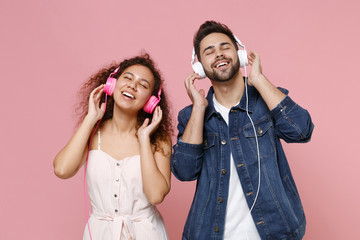 Joyful young couple two friends european guy african american girl in casual clothes isolated on pastel pink background. People lifestyle concept. Listen music with headphones, keeping eyes closed.