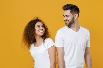 Fototapeta premium Smiling young couple friends european guy african american girl in white t-shirts isolated on yellow background. People lifestyle concept. Mock up copy space. Stand back to back looking at each other.