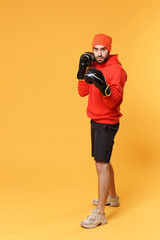 Young bearded fitness sporty guy boxer sportsman in hat, hoodie, shorts in home gym isolated on yellow background. Workout sport motivation lifestyle concept. Doing boxing exercises in boxing gloves.