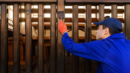 Contractor worker in blue overalls and protective gloves paints a fence
