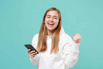 Happy young woman girl in casual white hoodie posing isolated on blue turquoise wall background studio portrait. People lifestyle concept. Mock up copy space. Using mobile phone, doing winner gesture.