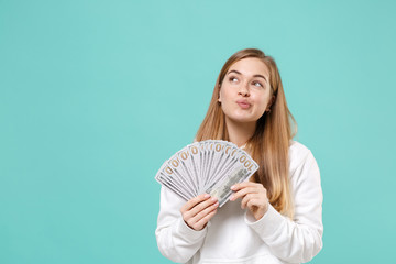 Pensive young woman girl in casual white hoodie posing isolated on blue turquoise wall background. People lifestyle concept. Mock up copy space. Hold fan of cash money in dollar banknotes, looking up.