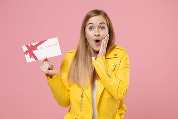 Shocked young woman girl in yellow leather jacket posing isolated on pastel pink background studio portrait. People lifestyle concept. Mock up copy space. Hold gift certificate, put hand on cheek.