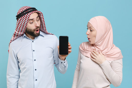 Shocked couple friends arabian muslim man wonam in keffiyeh kafiya ring igal agal hijab clothes isolated on blue background. People religious lifestyle concept. Hold mobile phone with empty screen.