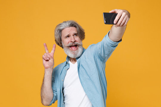 Funny elderly gray-haired mustache bearded man in casual blue shirt isolated on yellow background. People lifestyle concept. Mock up copy space. Doing selfie shot on mobile phone showing victory sign.