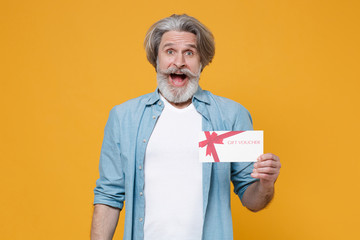 Surprised elderly gray-haired mustache bearded man in casual blue shirt posing isolated on yellow wall background studio portrait. People lifestyle concept. Mock up copy space. Hold gift certificate.