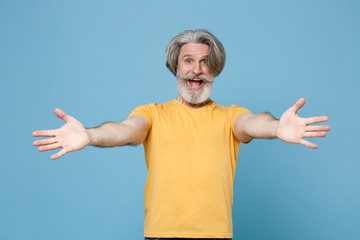 Excited elderly gray-haired mustache bearded man in casual yellow t-shirt posing isolated on blue background. People lifestyle concept. Mock up copy space. Standing with outstretched hands for hugs.