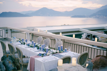 Wedding dinner table reception with lots thick blue candles against sunset sky over the mountains and sea in Budva, Montenegro. Old soft chairs on the restaurant's summer terrace, outside in open air.