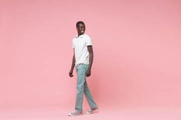 Fototapeta na wymiar Side view of smiling young african american man guy in white polo shirt, turquoise trousers posing isolated on pastel pink wall background. People lifestyle concept. Mock up copy space. Looking aside.