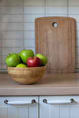 Wooden bowl with green apples and one red Apple on the kitchen table with a view of the kitchen table doors, cutting Board in the background