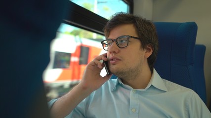 Handsome young businessman wearing a blue shirt and glasses is riding a train dialing a number and talking on his phone. Sunny summer day
