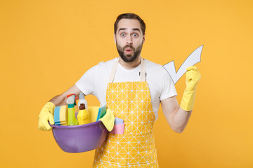 Shocked young man househusband in apron rubber gloves hold basin detergent bottles washing...