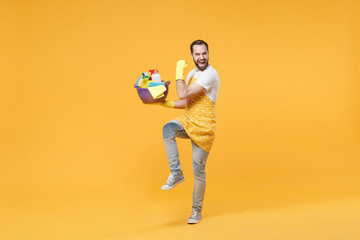 Joyful man househusband in apron gloves hold basin detergent bottles washing cleansers doing housework isolated on yellow background. Housekeeping concept. Mock up copy space. Doing winner gesture.