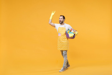 Joyful man househusband in apron gloves hold basin detergent bottles washing cleansers doing housework isolated on yellow background. Housekeeping concept. Waving greet with hand as notices someone.