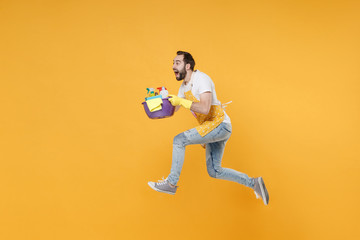 Fototapeta na wymiar Side view of crazy man househusband in apron rubber gloves hold basin detergent bottles washing cleansers doing housework isolated on yellow background. Housekeeping concept. Jumping like running.